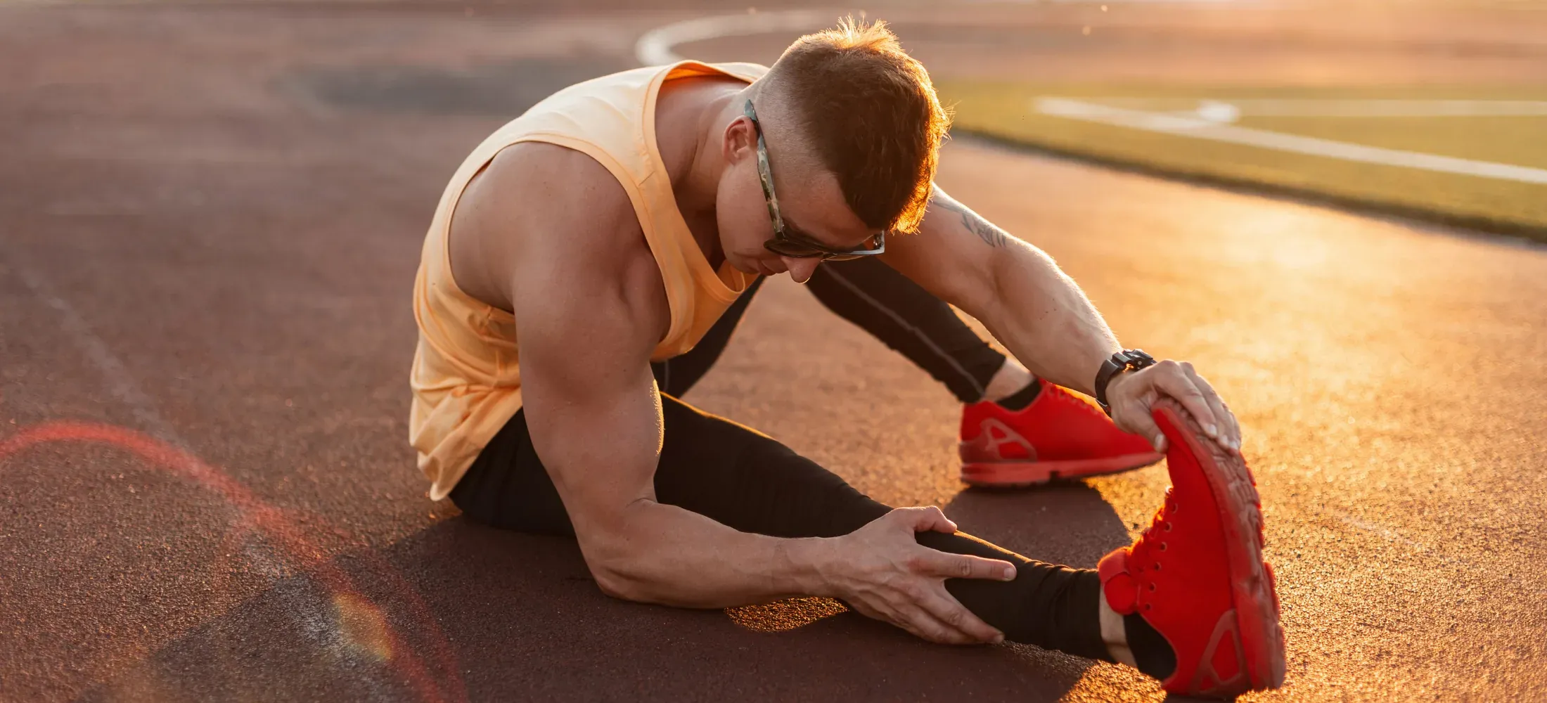 An athlete sitting on a running track at sunset, stretching forward towards the toes of one leg
