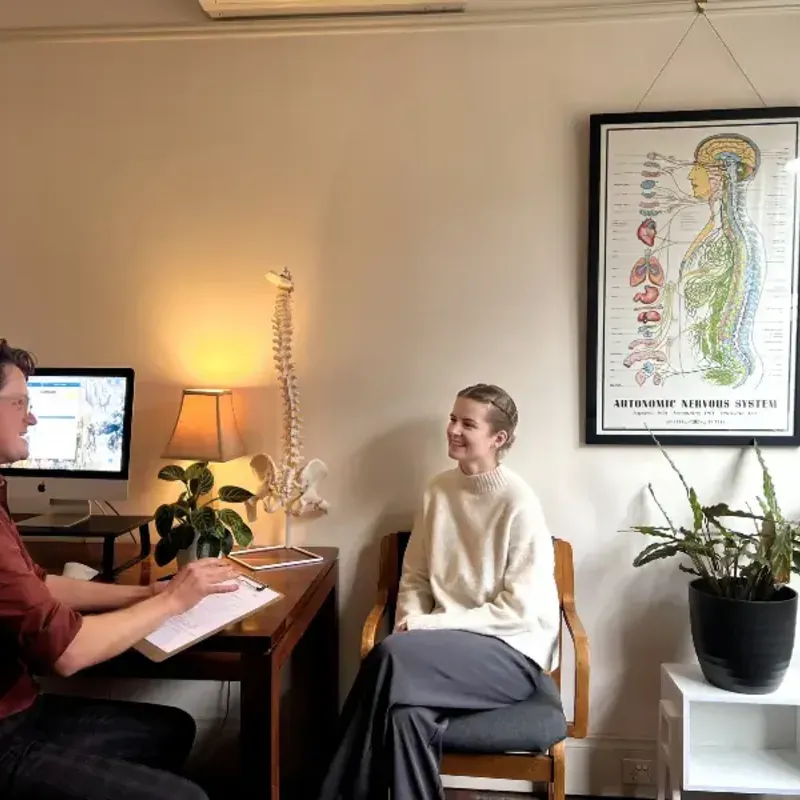 A chiropractor consulting with a patient beside an anatomical nervous system poster