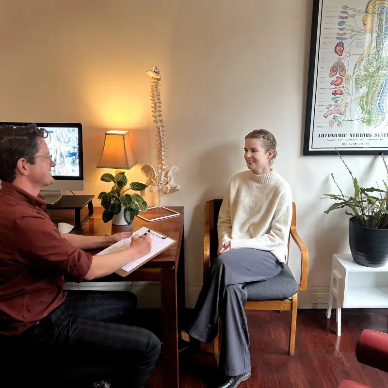 A practitioner making notes at a desk while talking with a seated patient in a consulting room at Brunswick Health, a spine model on the desk between them