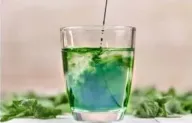 A green alkalising drink being stirred in a glass, with fresh leaves alongside