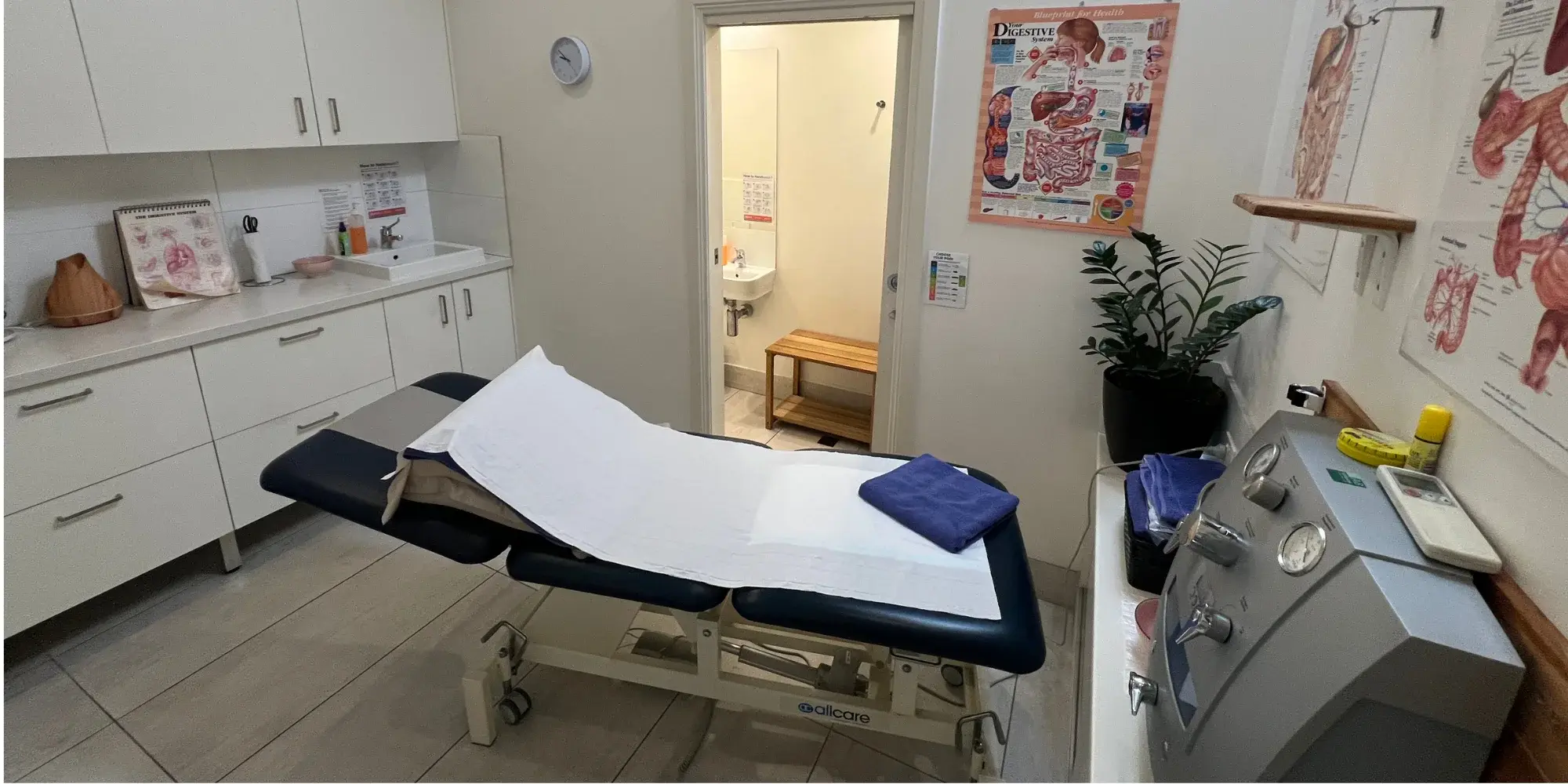A colonic irrigation treatment room at Brunswick Health — a draped table, the closed-system machine alongside it, and the room's own ensuite through an open door