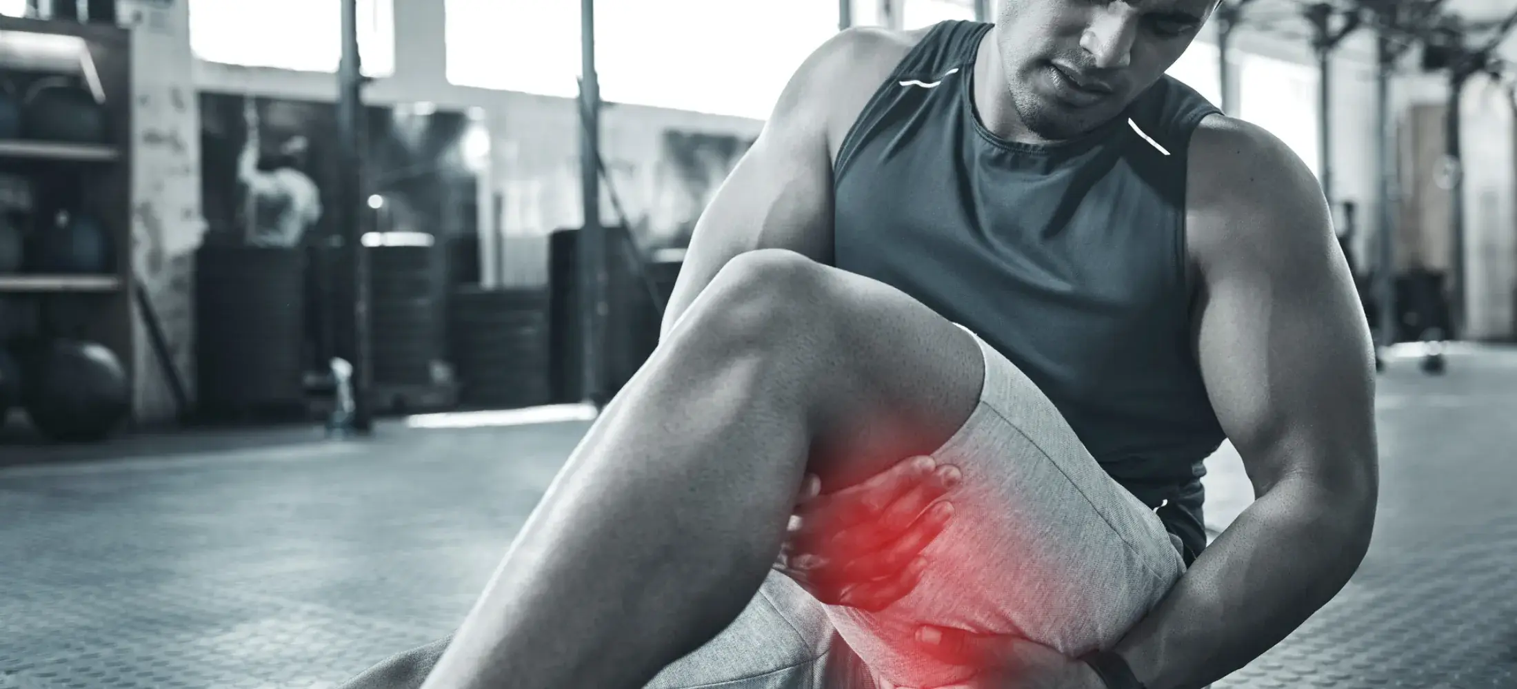 A man sitting on a gym floor gripping the back of his thigh, the hamstring highlighted