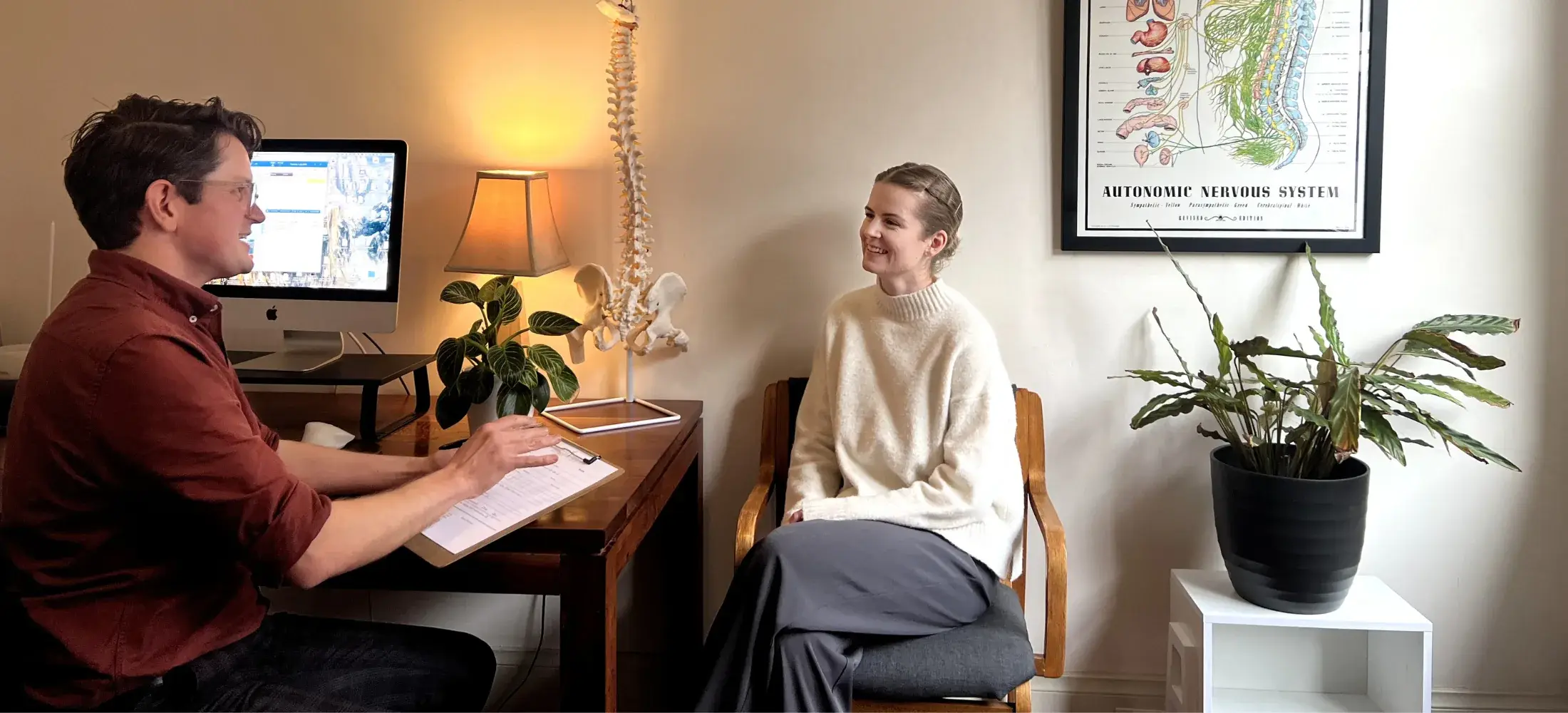 A practitioner with a clipboard talking with a seated patient in a consulting room at the clinic