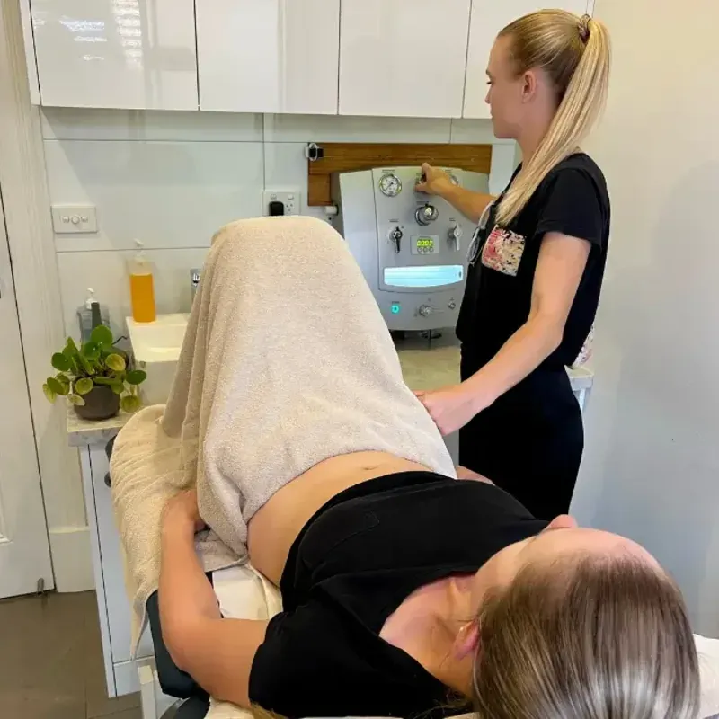 A colonic hydrotherapist beside a patient on the table during a session, the machine behind her