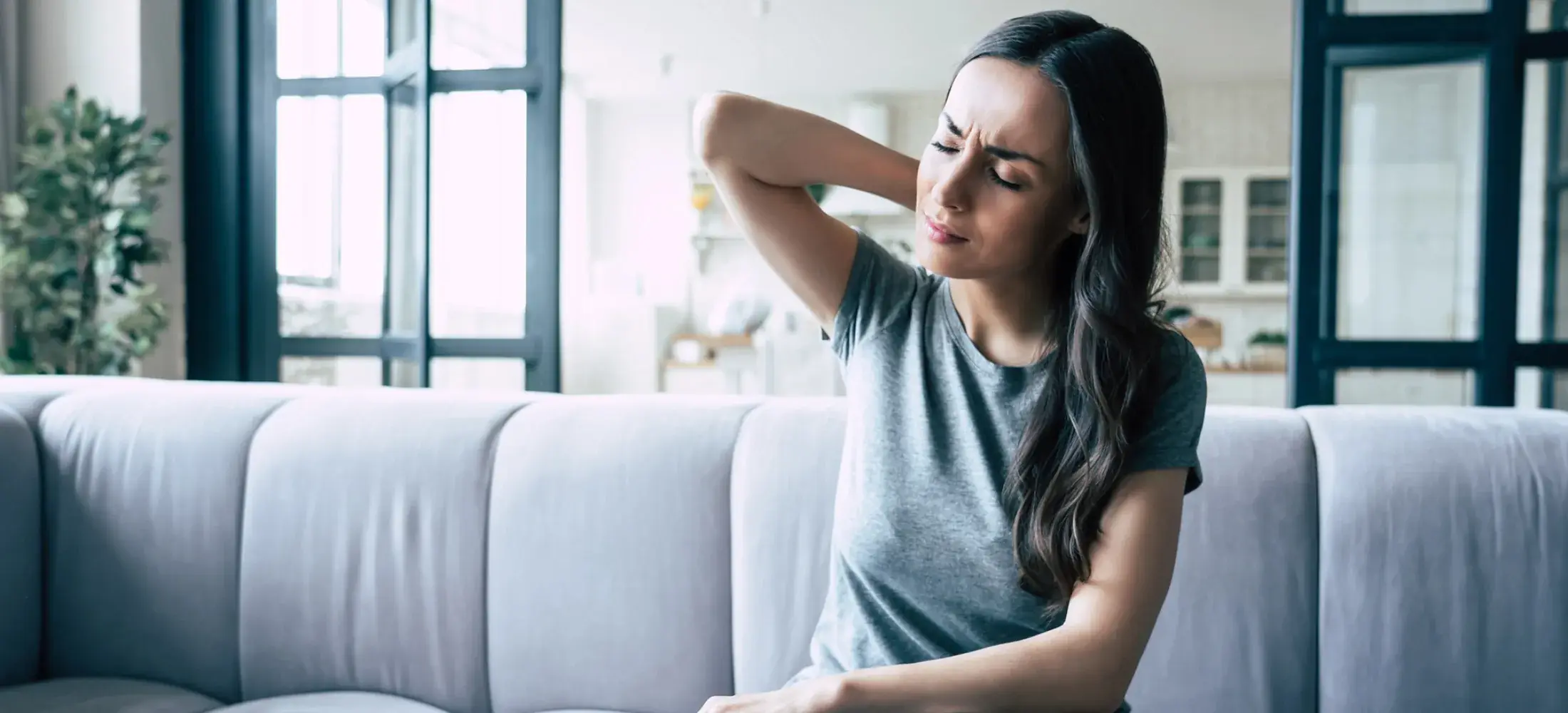 A woman on a sofa reaching a hand to the back of her neck and tilting her head to one side