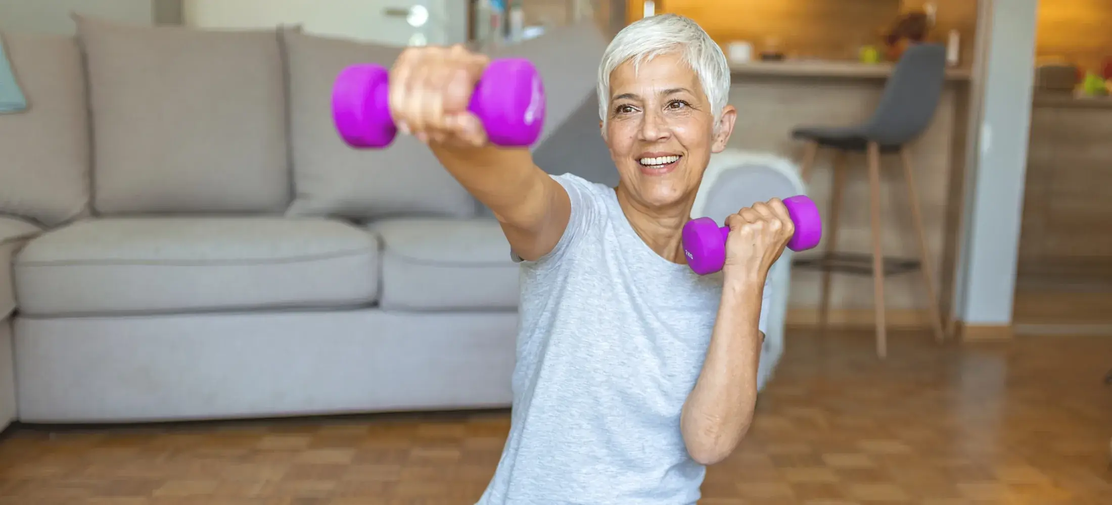 An older woman exercising at home with a dumbbell in each hand, one arm extended in front of her