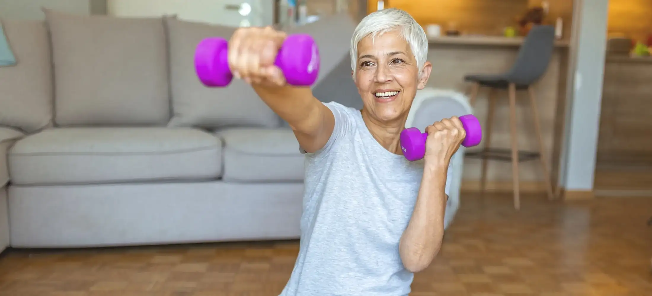 An older woman exercising at home with a dumbbell in each hand, one arm extended in front of her