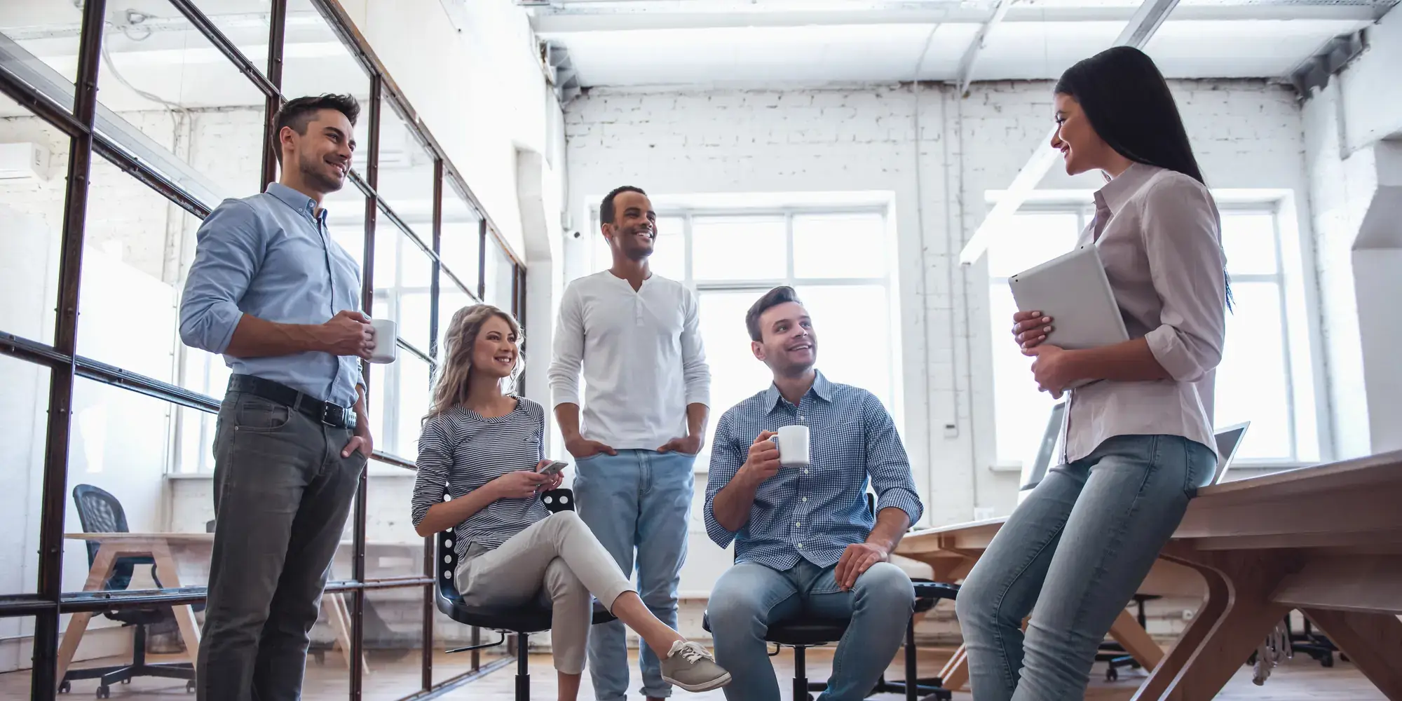 Five colleagues standing and sitting together with coffee in a bright open-plan workspace