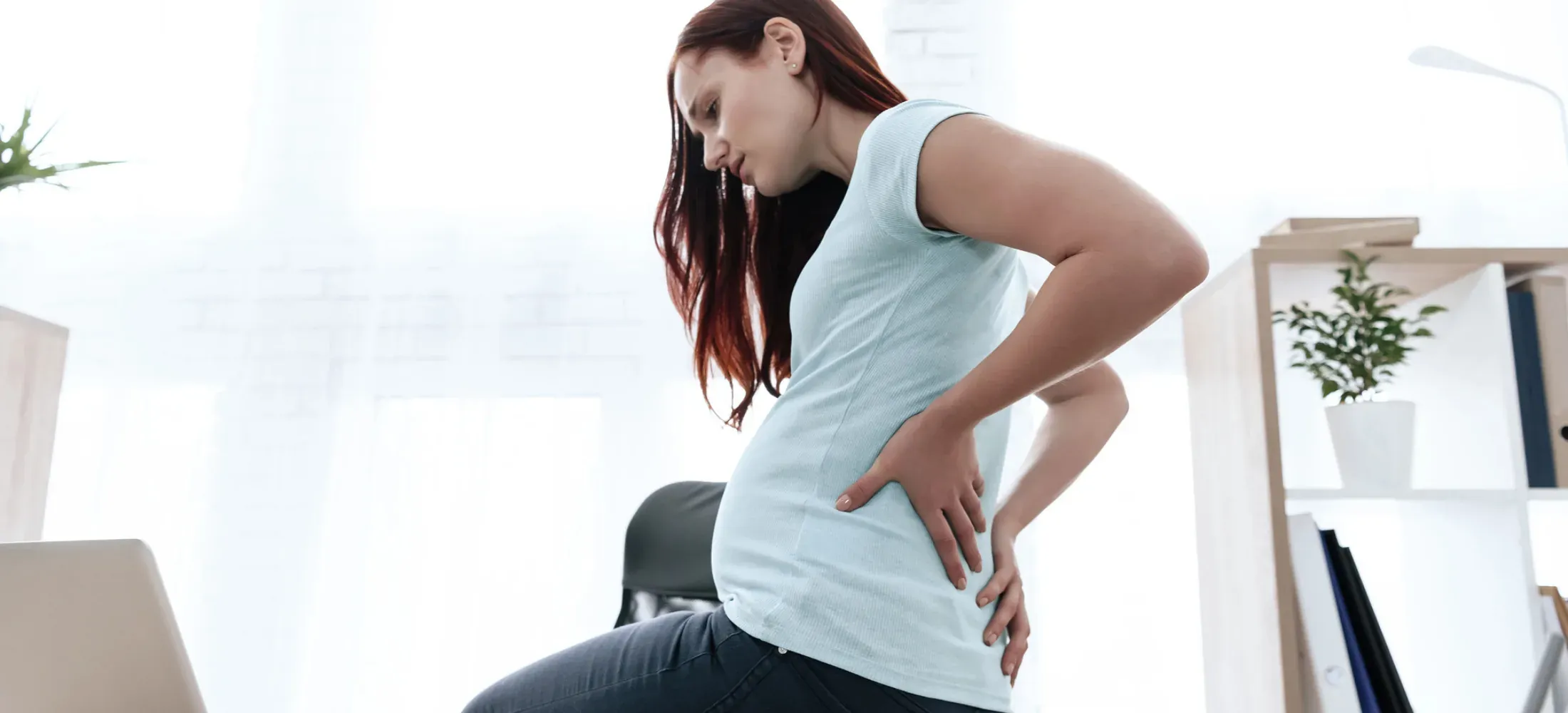A pregnant woman standing at a desk with a hand pressed to her lower back