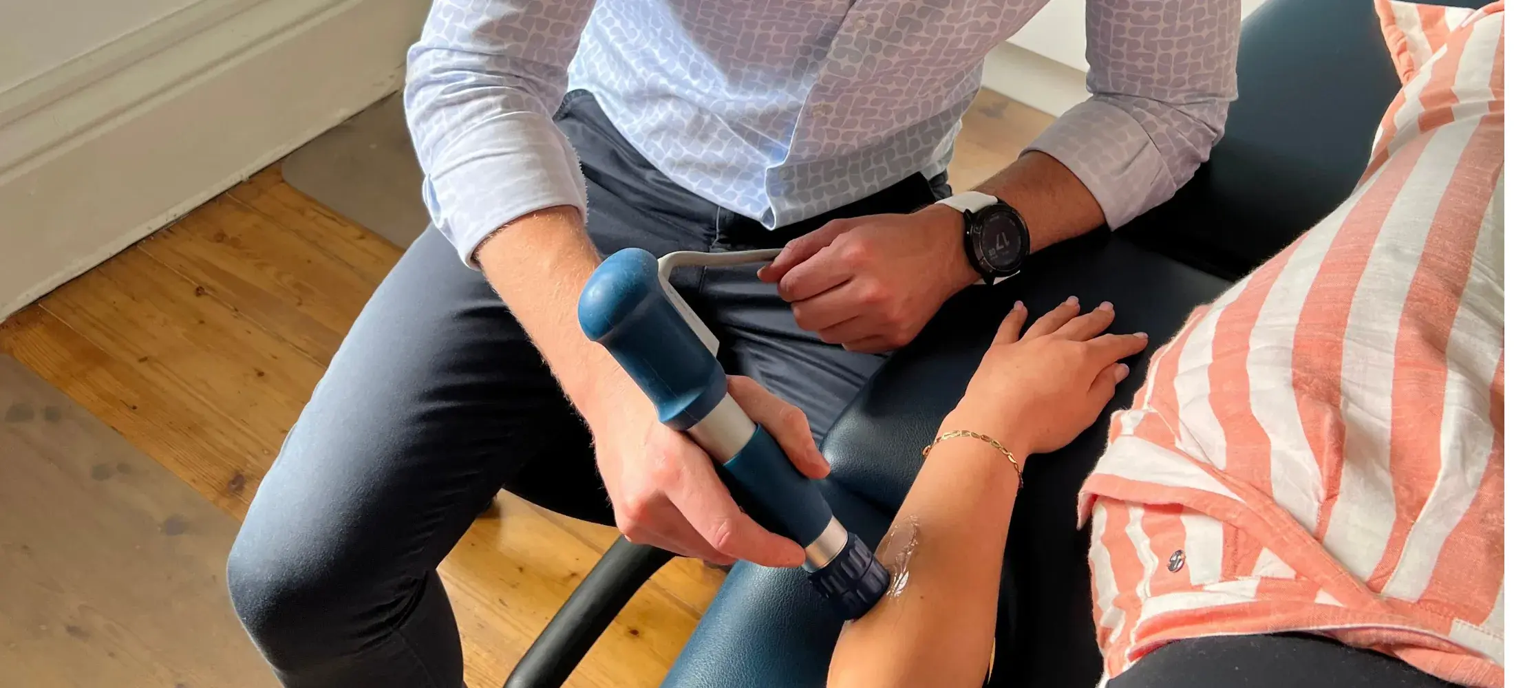 A practitioner applying a shockwave therapy handpiece to a patient's forearm at the clinic