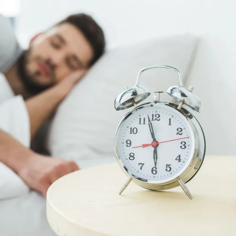 A man asleep in bed beside an alarm clock on the bedside table