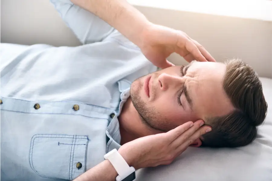 Young man lying on a bed, holding his temples and showing signs of pain, experiencing TMJ pain