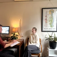 Chiropractor consulting with a female patient in a cozy office, with a detailed anatomical poster of the autonomic nervous system in the background.