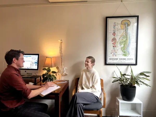 Chiropractor consulting with a female patient in a cozy office, with a detailed anatomical poster of the autonomic nervous system in the background.