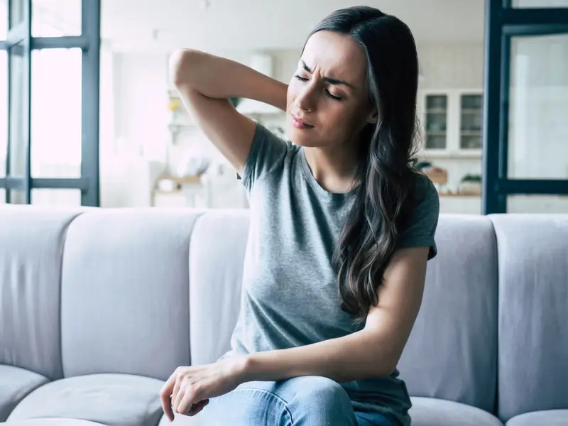 Woman holding her neck in pain while sitting on a couch at home contemplating seeing a Chiropractor or physiotherapist for Neck and shoulder pain treatment.