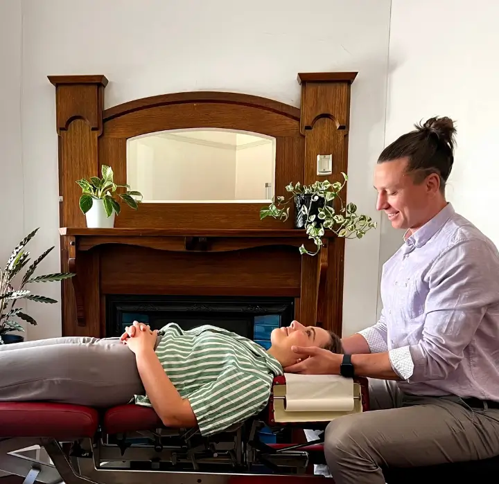 A melbourne brunswick chiropractor adjusts a patient's neck while she lies on a chiropractic table in a cozy room with a fireplace and plants.healthcare services.