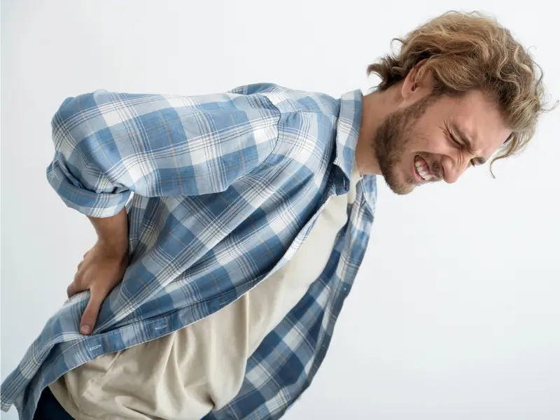 Man in a blue plaid shirt bent over in pain, clutching his lower back with one hand, visibly distressed, suggesting severe back pain or sciatica