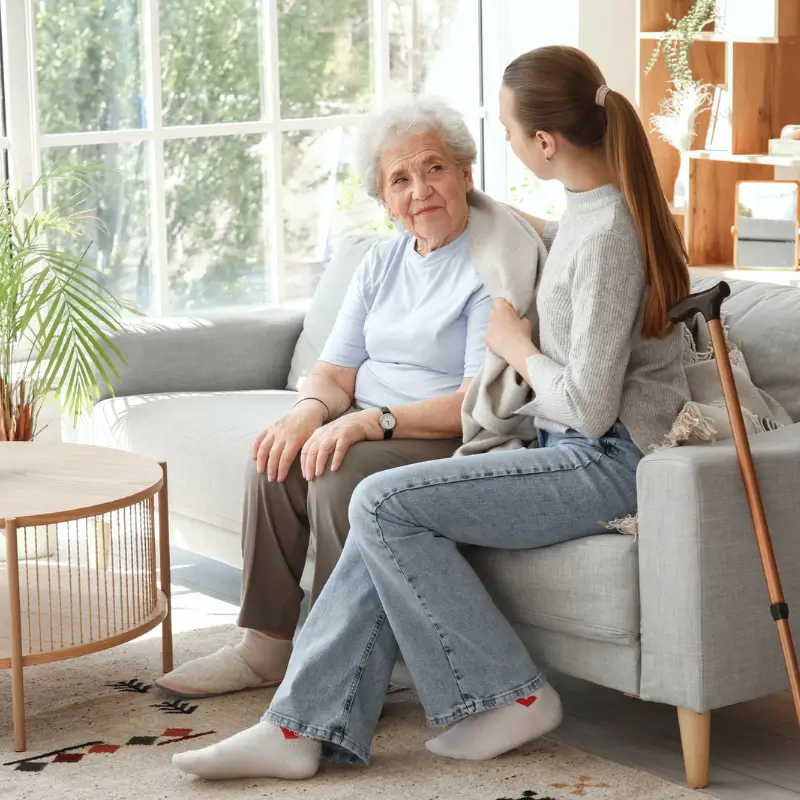 An elderly woman sitting on a sofa with her grand daughter in a brightly lit living room discussing the possibility of Aged Care Health Services such as Physiotherapy, Chiropractic, Massage, Podiatry, Osteopathy, Acupuncture to keep her at home as long as possible