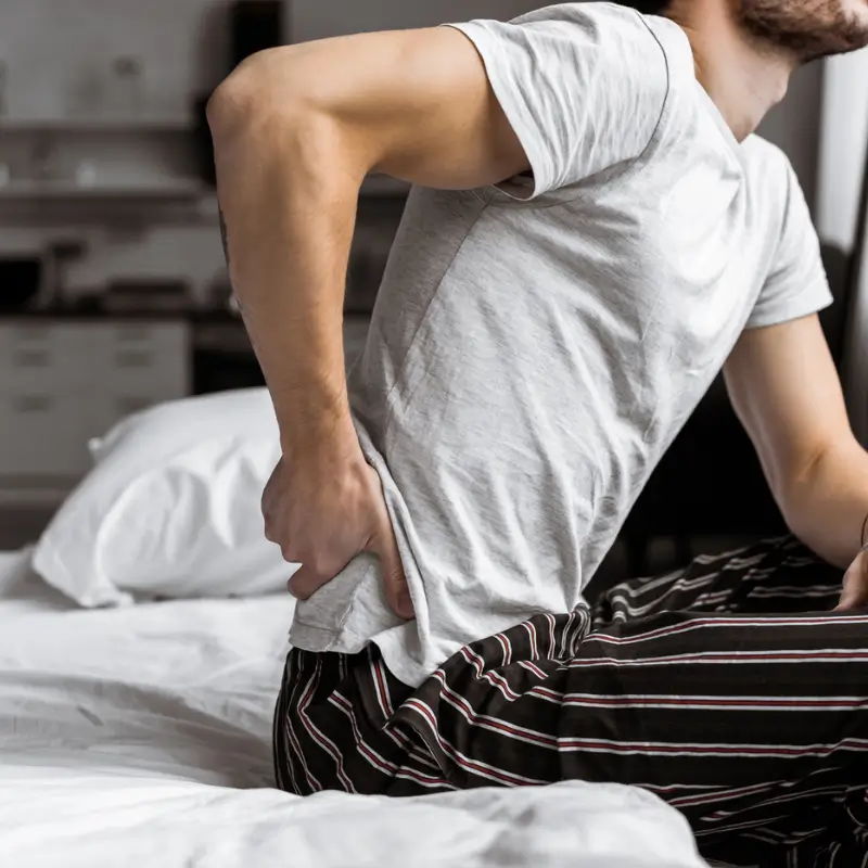 Man holding his lower back in pain while sitting on a bed, indicating discomfort from low back pain