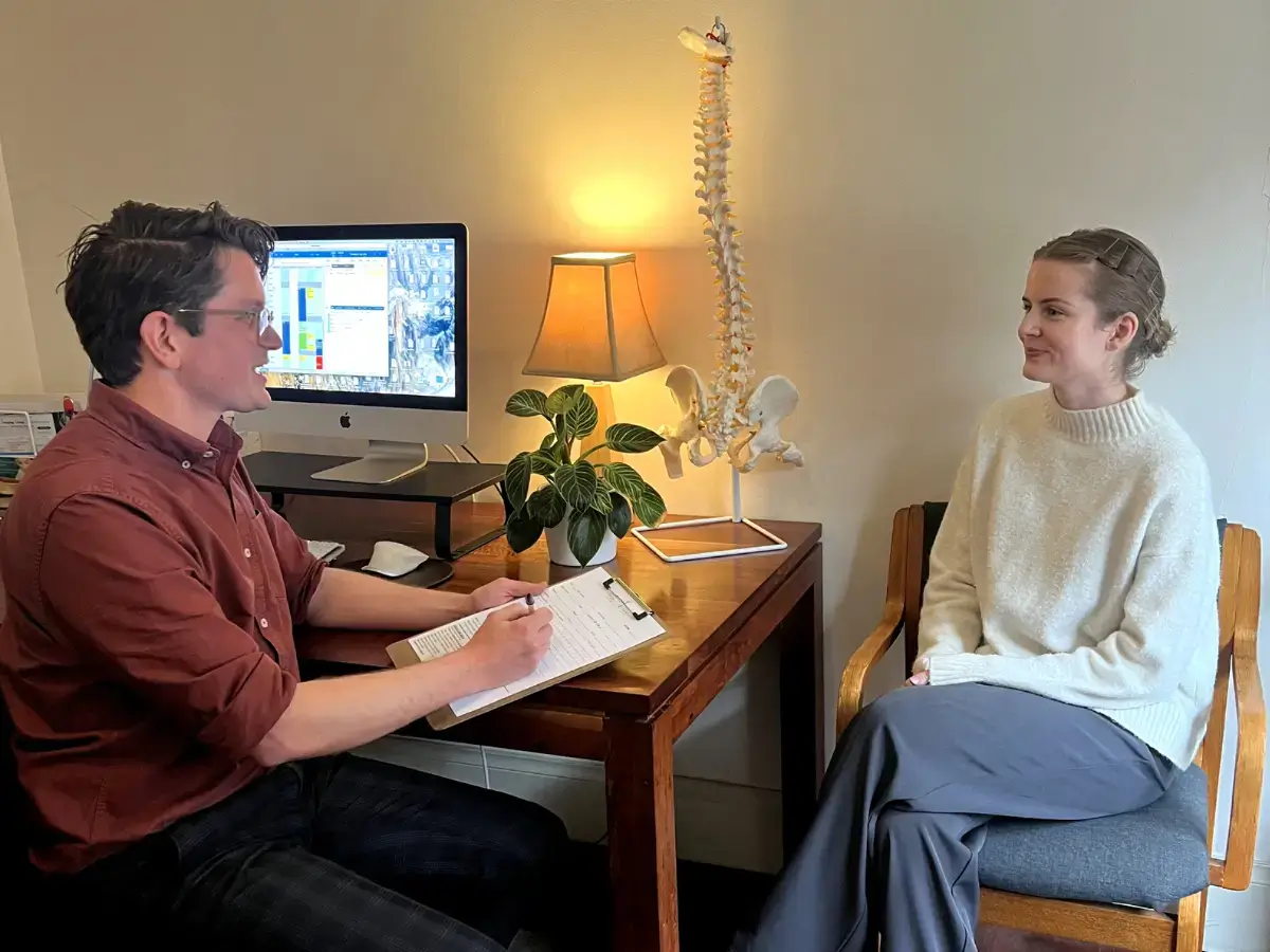 Chiropractor consulting with a female patient in a cozy office, with a detailed anatomical poster of the autonomic nervous system in the background.